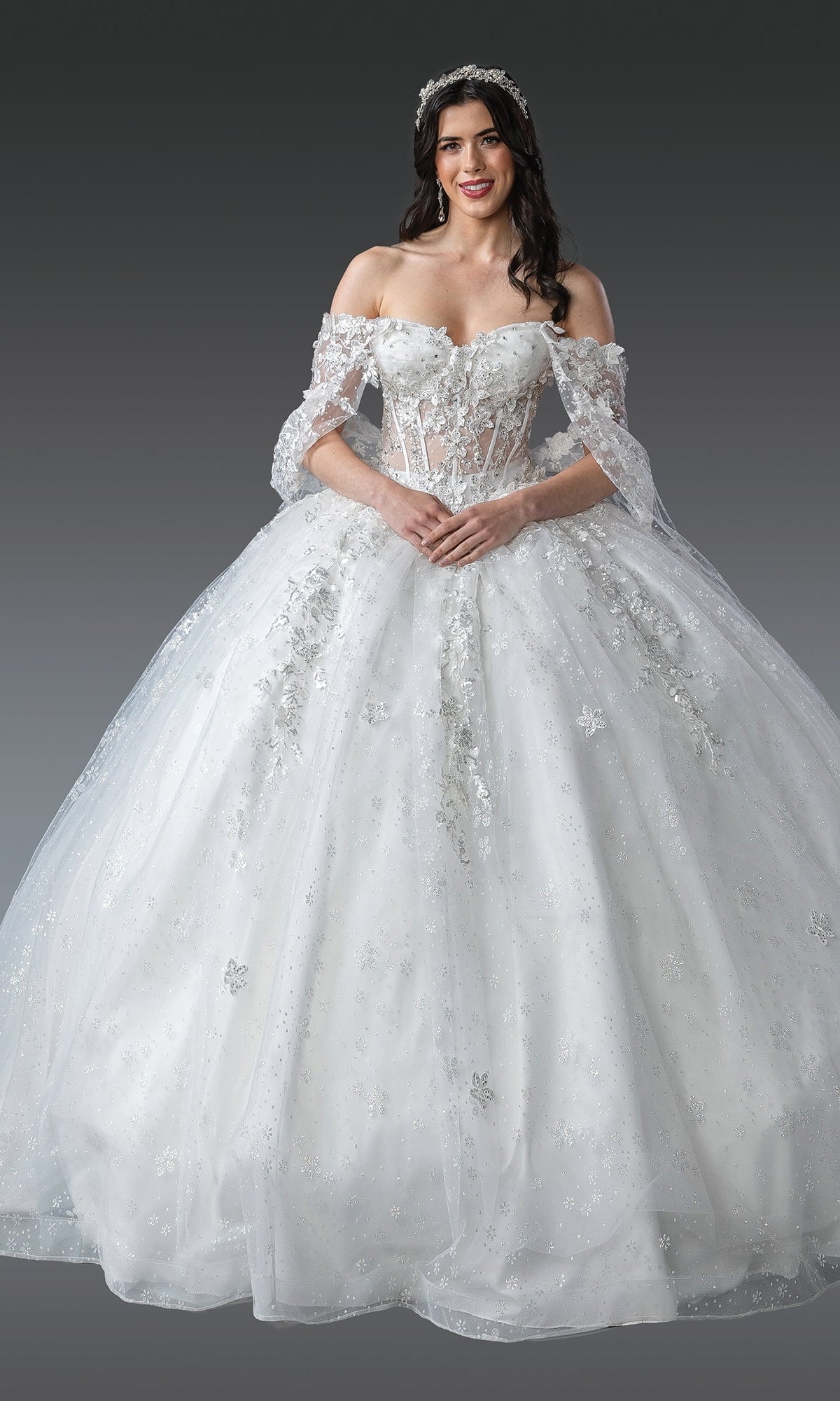A woman in the Quinceanera Dress 1970 By Dancing Queen, an off-the-shoulder white gown with 3D flowers and a full tulle ball skirt, wears a sparkling headpiece while standing against a plain gray background.